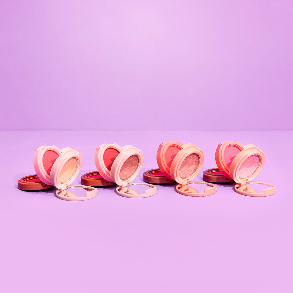 Dúo rubor e iluminador corazón compacto abierto mostrando rubor corazón e iluminador circular shimmer en múltiples tonos sobre fondo lila