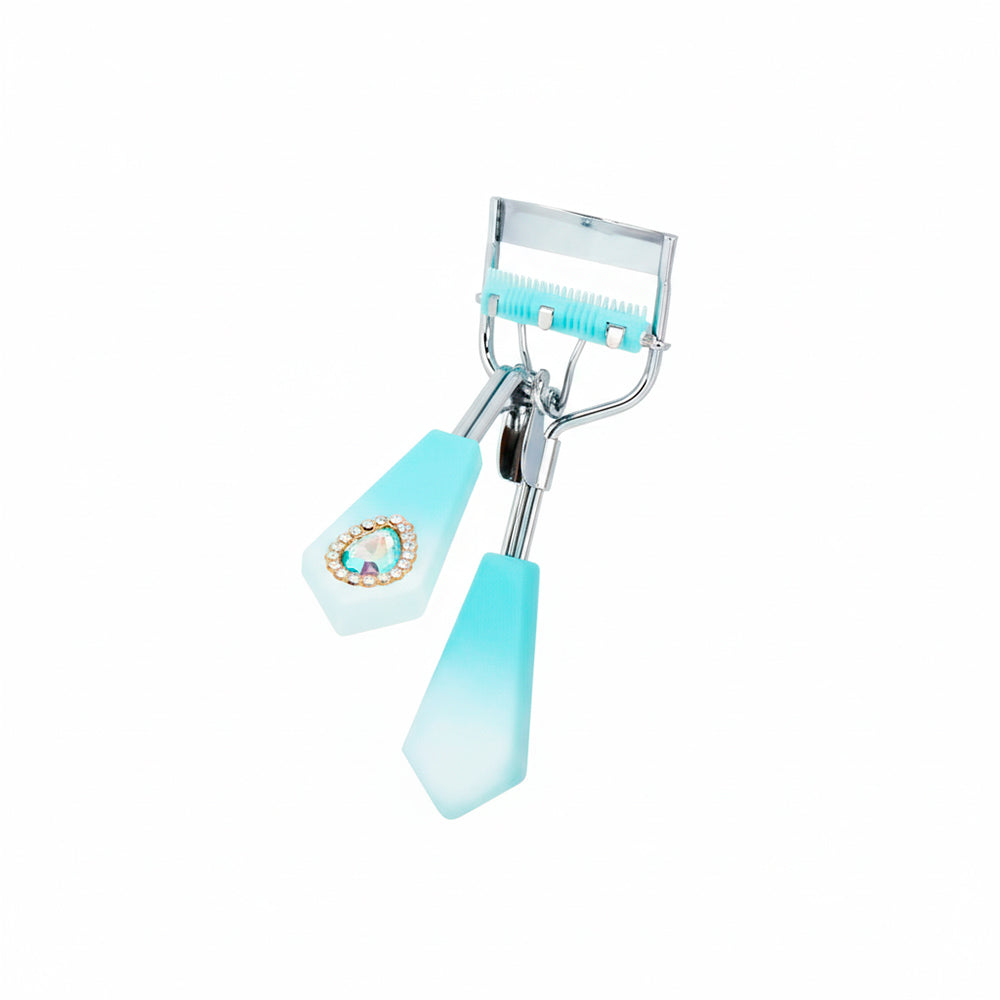 Rizador de pestañas Strass Heart Curl con mango facetado celeste degradé, detalle corazón strass y cerdas integradas celeste sobre fondo blanco