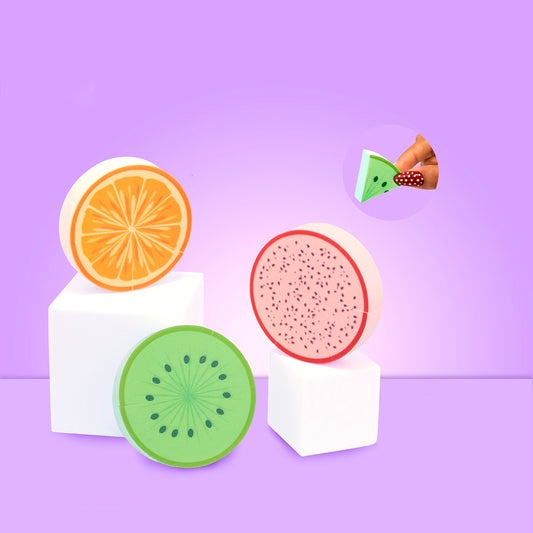 Beauty Blender Diseño Frutas – Tres esponjas redondas con diseño de naranja, kiwi y pitahaya sobre pedestales blancos, fondo lila
