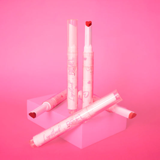 Labial en barra Reve'l Professional punta corazón ultra matte con empaque rosa estampado de corazones, tonos desde rosa suave hasta rojo vibrante, sobre fondo rosa