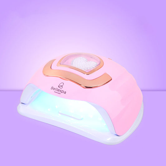 Lámpara UV LED para uñas Bellespa Cosmetics con ribete rose gold, panel LED en forma de corazón y base blanca con LEDs activos, sobre fondo lila degradado