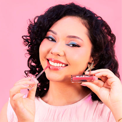 Modelo aplicando lip tint Reve'l Vinyl Ink en tono rosa coral con acabado glossy, look de maquillaje con delineado azul, sobre fondo rosa

