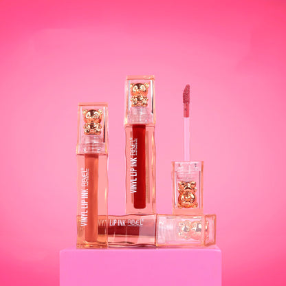 Lip tint Reve'l Vinyl Ink con tapa osito dorado, tonos nude coral y rojo intenso con aplicador aterciopelado, sobre fondo rosa