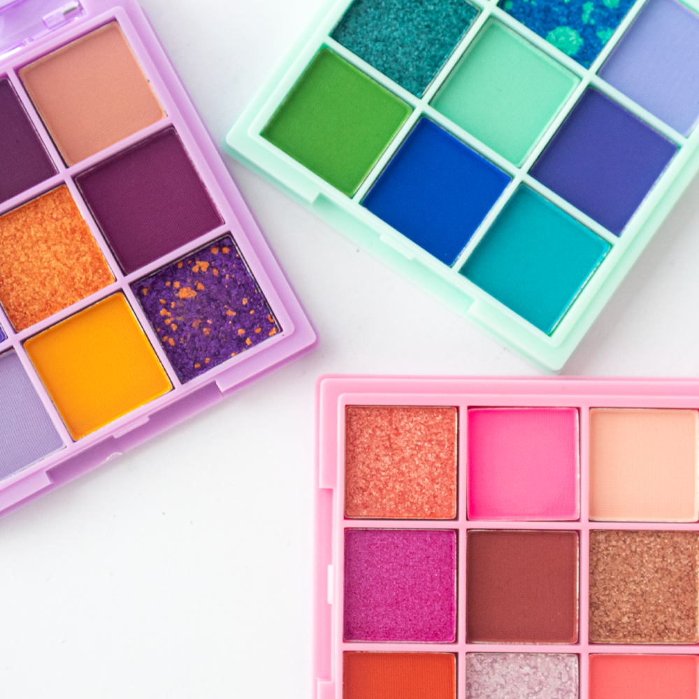 Detalle flat lay de las tres variantes de la Paleta de Sombras Diamond 9 Tonos Reve'l Professional con tonos púrpura, naranja y amarillo, verde y azul teal, y rosa coral y marrón con acabados mate y glitter