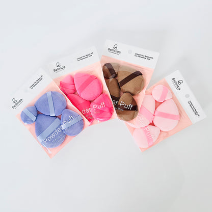 Set de 4 powder puff de terciopelo Bellespa Accesorios con cinta de agarre en colores lila fucsia marrón y rosa para aplicar polvo y base sobre fondo blanco